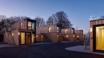 Un petit village de petites maisons habillées de bois et dotées de grandes fenêtres, construites à partir de conteneurs d’expédition recyclés, empilées les unes sur les autres. C’est le crépuscule et des lumières aux couleurs chaudes éclairent les intérieurs. (Photo : Stefan Hohloch pour Containerwerk) 