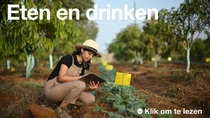 Een vrouwelijke landbouwer die een overall en een hoed draagt, houdt een tablet vast en onderzoekt planten in een veld op een zonnige dag. De tekst op de afbeelding luidt: "Eten en drinken."