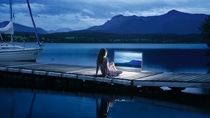A woman sits on a dock by the water and looks at a screen.