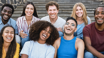 A diverse group of people sit together on outdoor steps, smiling and laughing at the camera, representing inclusion, connection, and a sense of belonging.