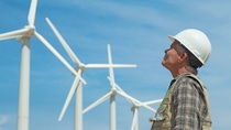 A man in a construction helmet looking up at several wind turbines.