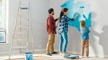 Family Time Together with Small Daughter. Young Father and Mother Showing Their Child How to Paint a Wall with a Roller. Paint Color is Light Blue. Room at Home is Prepared for Renovations. Family Time Together with Small Daughter. Young Father and Mother Showing Their Child How to Paint a Wall with a Roller. Paint Color is Light Blue. Room at Home is Prepared for Renovations.