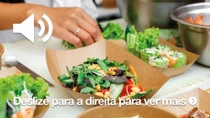 Vários itens alimentícios em recipientes de papel marrom. Uma mão é vista preparando a comida. Em segundo plano, há tigelas de metal contendo ingredientes adicionais.