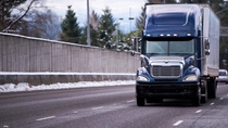 Dark blue big rig semi truck fleet with shiny chrome details and dry van semi trailer transporting industrial cargo on winter snowy road with concrete fence and snow on the shoulder. Dark blue big rig semi truck fleet with shiny chrome details and dry van semi trailer transporting industrial cargo on winter snowy road with concrete fence and snow on the shoulder.