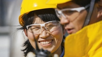 Close-up of two workers wearing yellow safety helmets and protective glasses, collaborating outdoors; one smiles while looking at a component or tool, the other focuses on the task in the foreground.