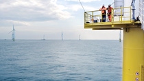 Eye level view of a yellow platform at the base of a wind tubine, with site workers in orange overalls leaning on the railing to take a break and look at the view. Photo: Vattenfall Eye level view of a yellow platform at the base of a wind tubine, with site workers in orange overalls leaning on the railing to take a break and look at the view. Photo: Vattenfall
