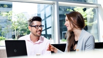 Two people sit together with a laptop in a bright office, smiling and engaged in a friendly interview conversation.