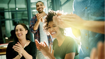 A cheerful group of colleagues applaud and laugh in a modern office as sunlight streams through the windows.