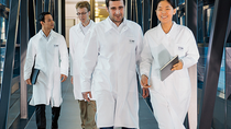 Eine Gruppe von Wissenschaftler:innen in Laborkitteln geht durch einen modernen Gang. Eine Gruppe von Wissenschaftler:innen in Laborkitteln geht durch einen modernen Gang.