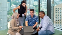 A diverse team of four colleagues collaborating around a laptop in a modern office with a city skyline in the background.