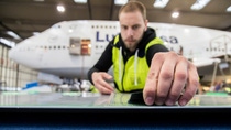 A young male technician wearing a high-visibility jacket stands in an aircraft hangar with a Lufthansa aircraft in the background and carefully applies sharkskin film to a flat surface. A young male technician wearing a high-visibility jacket stands in an aircraft hangar with a Lufthansa aircraft in the background and carefully applies sharkskin film to a flat surface.