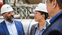 Three colleagues wearing white safety helmets and protective glasses talk and smile inside an industrial plant, with pipes and equipment visible in the background.