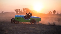 Partly silhouetted against the sunset, racing driver Hernan Bradas and his co-driver wave as they drive the green and blue eBaquet, an electric car built in the style of a famous 1940s racing car, along a dusty track in Argentina. 