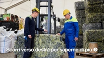 Due uomini in caschi gialli e tuta da lavoro sono in piedi accanto a un grande blocco pressato di rifiuti tessili.&nbsp;