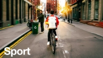 Een fietser met een rood jack en een witte rugzak, gemarkeerd met een gloeiend lichtringeffect, fietst bij zonsondergang door een stadsstraat en passeert op de achtergrond een persoon die aan het recyclen is. De tekst luidt: ‘Sport’.