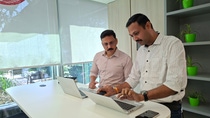 Two employees collaborate at a standing desk in a modern office, working on laptops and reviewing information together.