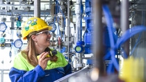 An employee wearing protective helmet, eyewear, and workwear inspects equipment in a chemical processing facility while holding a device and clipboard.