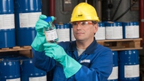 BASF employee in blue overalls, green gloves and yellow hard hat inspecting a bottle of chemical 