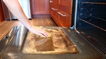 A close-up image of an open oven glass door with dirty greasy stains on it covered with special chemical foam for oven cleaning and woman's hand holding a cleaning sponge cleaning this oven. Ceramic tile floor on the background. Modern kitchen interior. Dirty oven. Glass oven door is being cleaned. A close-up image of an open oven glass door with dirty greasy stains on it covered with special chemical foam for oven cleaning and woman's hand holding a cleaning sponge cleaning this oven. Ceramic tile floor on the background. Modern kitchen interior. Dirty oven. Glass oven door is being cleaned.