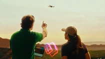 A male and female farmer are standing outdoors in South America, facing away from the camera, observing a drone flying in the sky. The man is holding a tablet and points towards the drone. The woman is looking at the drone. A light swirl hovers between them.