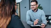 A friendly consultant in a gray sweater conducts a personal conversation with a customer in a modern office while taking notes on a clipboard.