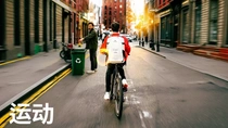 A cyclist with a red jacket and white backpack, marked with a glowing light-ring effect, rides down a city street at sunset, past a person recycling in the background. The text reads: "Sports."