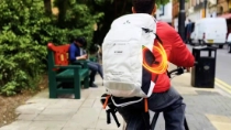 A person seen from behind on a bicycle wears a white backpack with VAUDE and BASF log. An orange light swirl appears on the backpack.