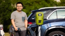 A man with an electric charger in his hand stands in front of electric cars at an electric charging station.