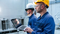 Two employees in an industrial facility use a laptop and smart glasses for equipment inspection.