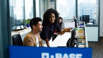 Colleagues in a modern office collaborate at a computer, with one person pointing at the screen while others observe and work at nearby desks.