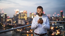 A portrait of businessman with smartphone standing against night London rooftop view panorama. Copy space.
