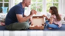Father and daughter sitting on a sofa eating pizza.