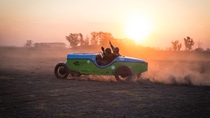 Partly silhouetted against the sunset, racing driver Hernan Bradas and his co-driver wave as they drive the green and blue eBaquet, an electric car built in the style of a famous 1940s racing car, along a dusty track in Argentina. 