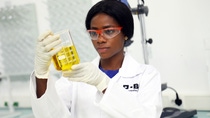 Laboratory employee wearing protective eyewear and gloves carefully examines a beaker containing a yellow liquid in a controlled lab environment.