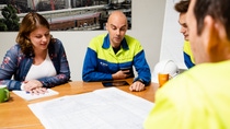Four employees, including technical staff in workwear, collaborate at a table reviewing technical drawings and project documents.