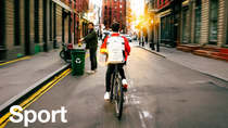 Ein Radfahrer in roter Jacke mit weißem Rucksack, der mit einem leuchtenden Lichtring-Effekt markiert ist, fährt in der Abenddämmerung eine Stadtstraße entlang, im Hintergrund eine Person beim Recycling. Der Text lautet: „Sports."
