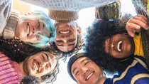 A diverse group of people in casual clothing lean together in a circle, smiling and looking down at the camera, representing connection, inclusion, and team spirit.