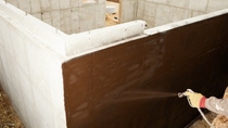 A worker is spraying waterproof sealant on a new home's concrete foundation. This area of the foundation will be below ground and the tar like material prevents water from contacting the concrete. Focus is on the wall in the area of the spray, the hand and sprayer are slightly soft. A worker is spraying waterproof sealant on a new home's concrete foundation. This area of the foundation will be below ground and the tar like material prevents water from contacting the concrete. Focus is on the wall in the area of the spray, the hand and sprayer are slightly soft.