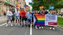 A diverse group of BASF employees walk together in a city street during a Pride parade, holding colorful signs and a rainbow-themed banner reading “Together for openness and respect,”.