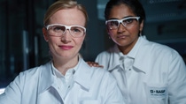 Two female scientists wearing white lab coats with the BASF logo and safety goggles stand in the laboratory and look into the camera.