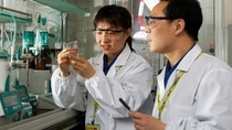 Two laboratory employees wearing protective eyewear and lab coats examine a liquid sample in a beaker, discussing results in a laboratory setting.