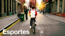 Um homem com mochila branca anda de bicicleta por uma rua da cidade ao pôr do sol, enquanto outro homem aparece ao fundo perto de uma lixeira de reciclagem. O texto diz: "Esportes."