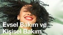 Siyah kıvırcık saçlı gülümseyen bir kadın. Resimdeki metinde şunlar yazıyor: “Evsel ve Kişisel Bakım.”