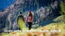 Un uomo e una donna attraversano a piedi un prato di montagna in una giornata di sole. Indossano abiti caldi da esterno.