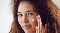 Portrait of woman with curly hair and freckles applying moisturizer to her face.