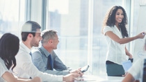 African American woman giving a presentation to her team. She is using a whiteboard with charts and graphs. She is talking. They are in an office boardroom at the table with laptop computers and paperwork. She is smiling. Men and women in the group.