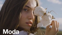 Una mujer con el cabello castaño largo sostiene una planta de algodón cerca de su cara. En el texto de la imagen pone: “Moda”
