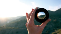 A hand is holding a lens in front of a mountain, focusing on the summit