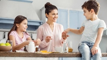 Happy mother. Attractive alert dark-haired young mother smiling and giving pills to her son sitting on the table and her daughter having breakfast Happy mother. Attractive alert dark-haired young mother smiling and giving pills to her son sitting on the table and her daughter having breakfast