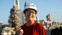 A BASF employee wearing a safety helmet and protective glasses stands at an industrial site, smiling and holding a walkie-talkie, with production facilities in the background.